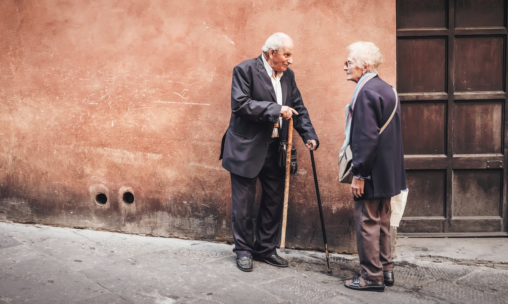 Two old men chatting while standing against a wall.